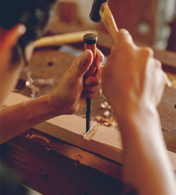 Strukturbau GmbH Oldenburg Eine Person benutzt einen Meißel und einen Hammer, um ein rechteckiges Loch in ein Stück Holz zu schnitzen, wobei sie sich auf die Holzbearbeitung an einer Werkbank konzentriert.