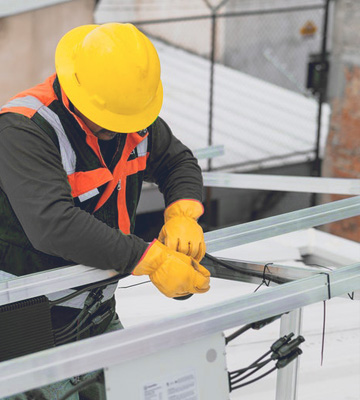 Strukturbau GmbH Oldenburg Ein Bauarbeiter, der einen gelben Schutzhelm, eine orangefarbene Sicherheitsweste und gelbe Handschuhe trägt, installiert Kabel auf einem Metalldach, wobei im Hintergrund Gebäude und ein Drahtzaun zu sehen sind.