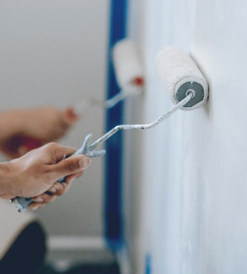 Strukturbau GmbH Oldenburg Zwei Personen verwenden Farbroller, um weiße Farbe auf eine Wand aufzutragen. Die Hände der einen Person sind im Fokus und halten die Walze nahe an der Wand, während die andere Walze etwas weiter entfernt ist.