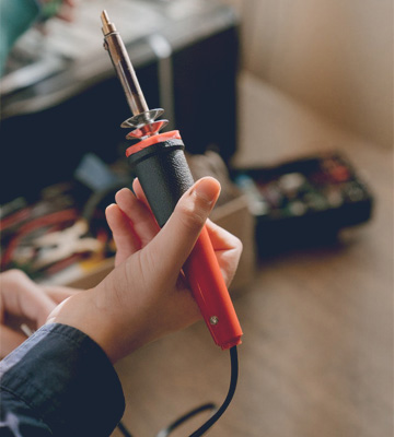 Strukturbau GmbH Oldenburg Eine Person hält einen rot-schwarzen Lötkolben in der Hand, im unscharfen Hintergrund sind Werkzeuge und Geräte auf einem Tisch zu sehen.