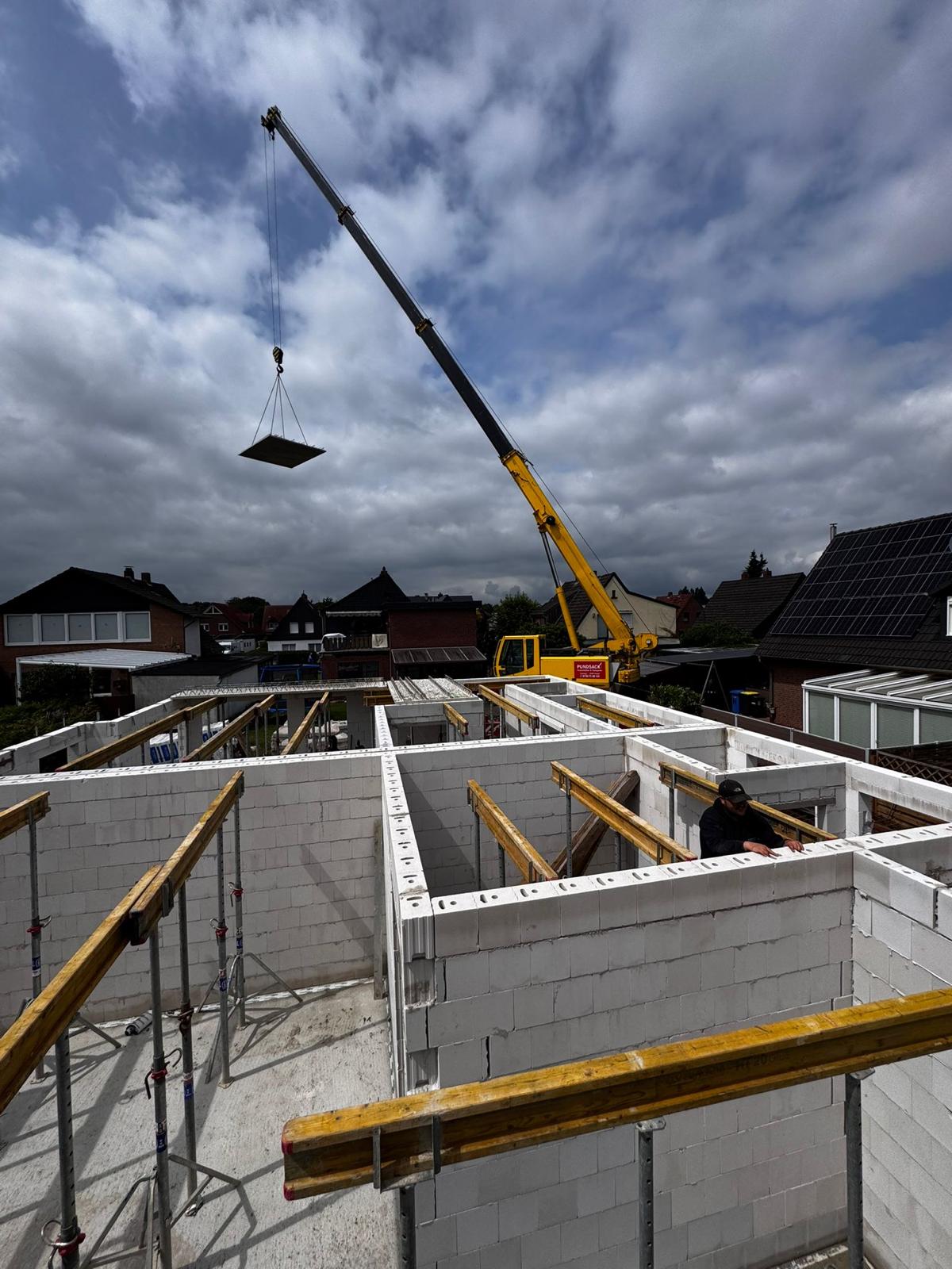 Strukturbau GmbH Oldenburg Ein gelber Kran eines Oldenburger Bauunternehmens hebt Baumaterial über ein teilweise gebautes Haus mit weißen Ziegelwänden; Gerüste und Balken sind sichtbar, und ein Arbeiter steht im Inneren. Wohnhäuser und ein bewölkter Himmel vervollständigen die Szene.