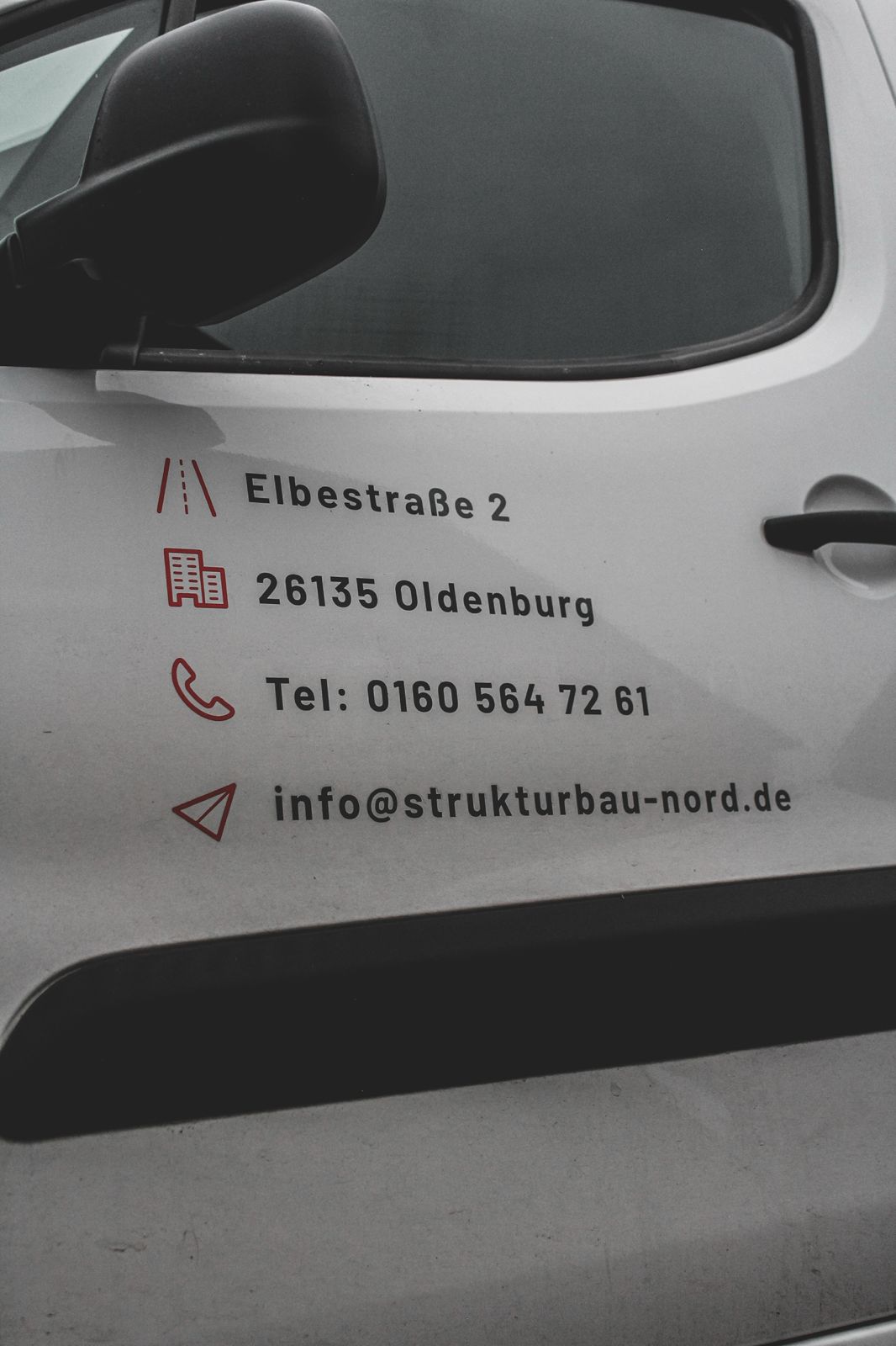 Strukturbau GmbH Oldenburg Weiße Fahrzeugtür mit Geschäftskontaktdaten für Bauunternehmen Oldenburg: Adresse Elbestraße 2, 26135 Oldenburg; Telefon 0160 564 72 61; E-Mail info@strukturbau-nord.de. Rote Symbole markieren die Adresse, das Telefon und die E-Mail.