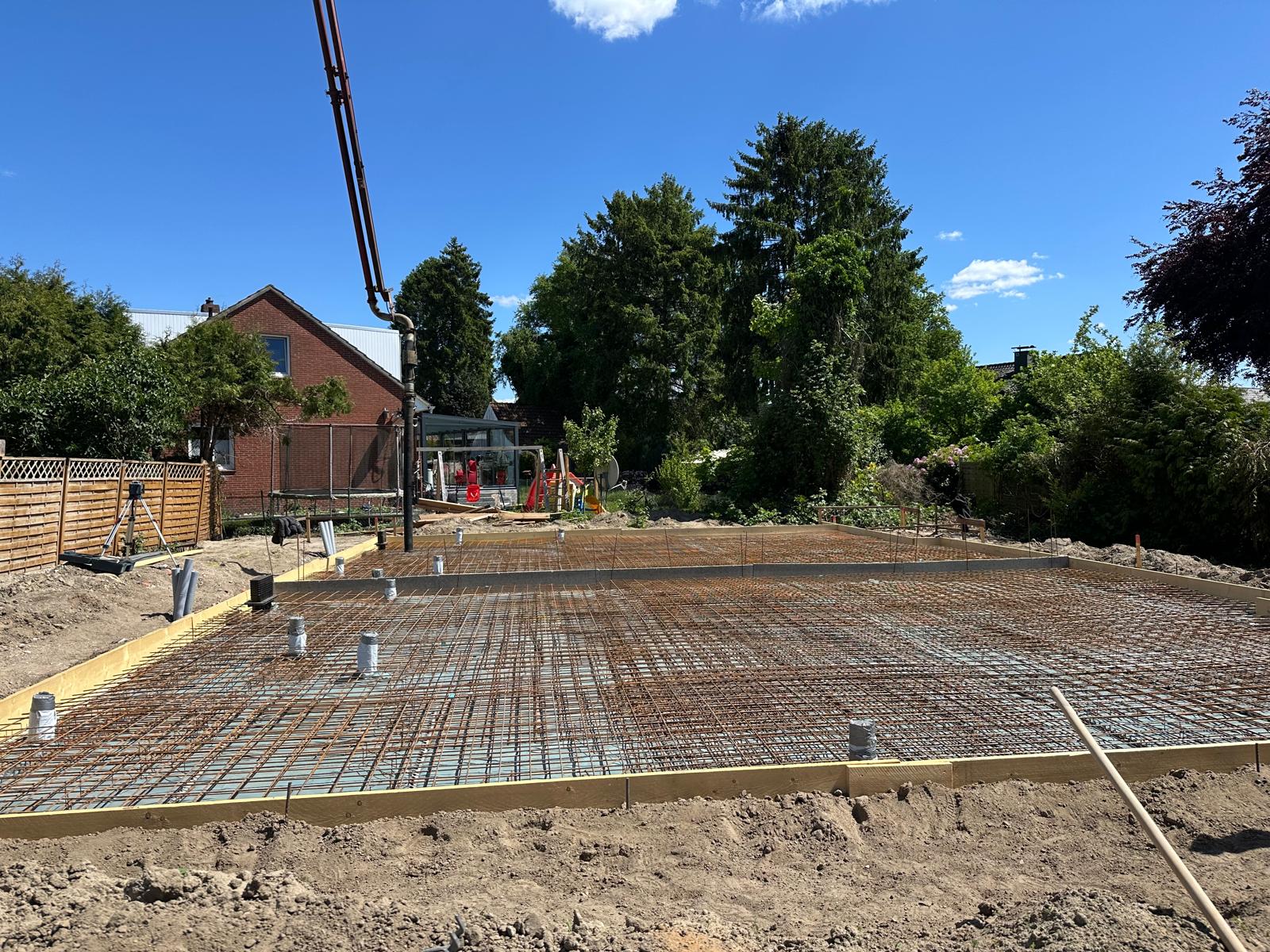 Strukturbau GmbH Oldenburg Eine Baustelle mit einer Fundamentplatte aus Beton, die mit Bewehrungsstahl verstärkt wird. Aus der Platte ragen Rohre heraus, und im Hintergrund sind Bäume, Häuser und ein strahlend blauer Himmel zu sehen.