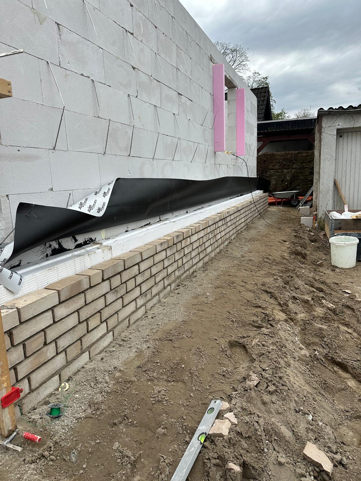 Strukturbau GmbH Oldenburg Eine teilweise errichtete Ziegelmauer grenzt an ein weißes Gebäude, in dem Isolierung und schwarzes Abdichtungsmaterial sichtbar sind. Bauwerkzeuge, Eimer und Schmutz sind auf dem Boden neben dem Gebäude verstreut.