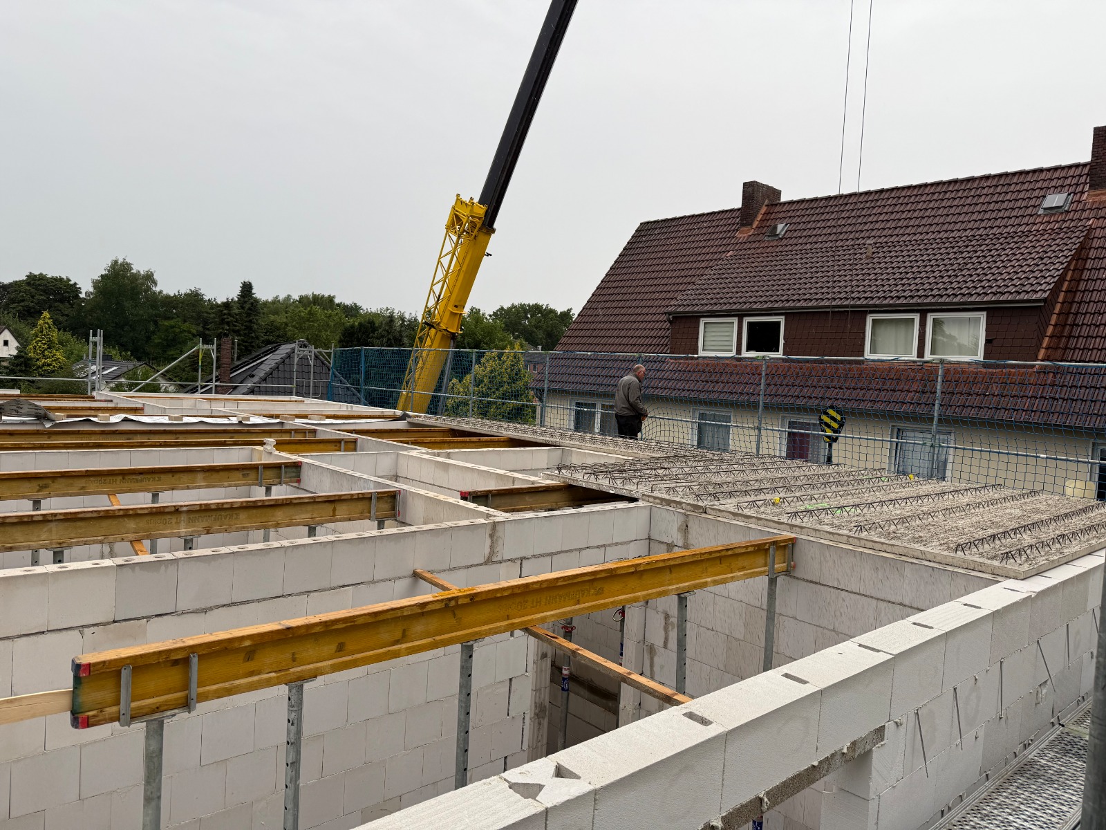 Strukturbau GmbH Oldenburg Eine Baustelle mit weißen Ziegelwänden, Holzbalken und einem Kran, der Material anhebt. Ein Arbeiter steht auf einer Betonplatte, und im Hintergrund ist ein Haus mit einem braunen Ziegeldach zu sehen.