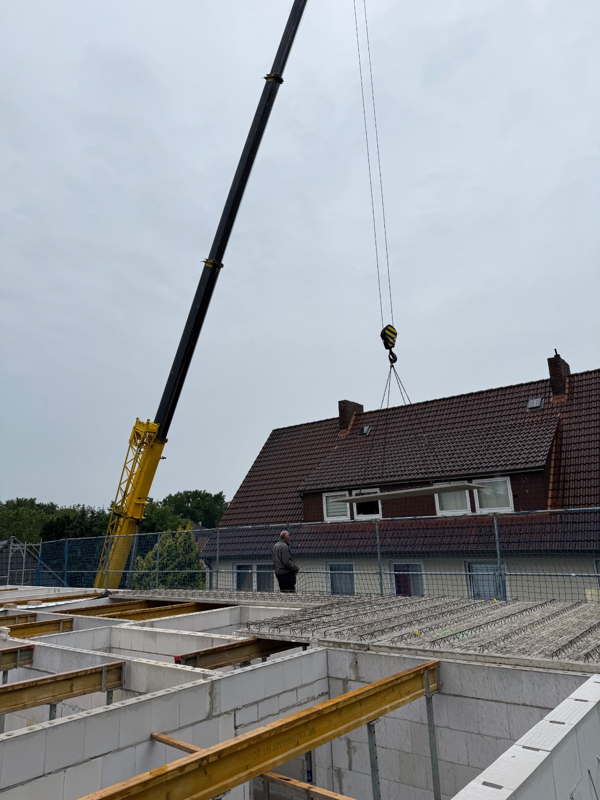 Strukturbau GmbH Oldenburg Ein großer gelber Kran hebt Materialien in der Nähe eines Wohnhauses mit einem Schrägdach. Eine Person steht unten an einem unfertigen Gebäude mit freiliegenden Balken und weißen Ziegelwänden. Der Himmel ist bedeckt.