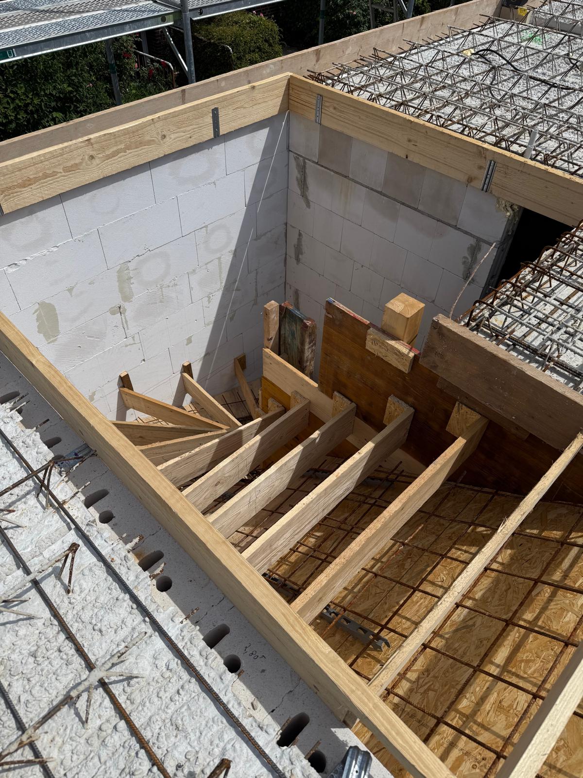 Strukturbau GmbH Oldenburg Holztreppenschalung im Bau auf einer Baustelle, umgeben von Betonblöcken und Bewehrungsstäben, mit teilweise errichteten Wänden und noch nicht installierten Treppen.