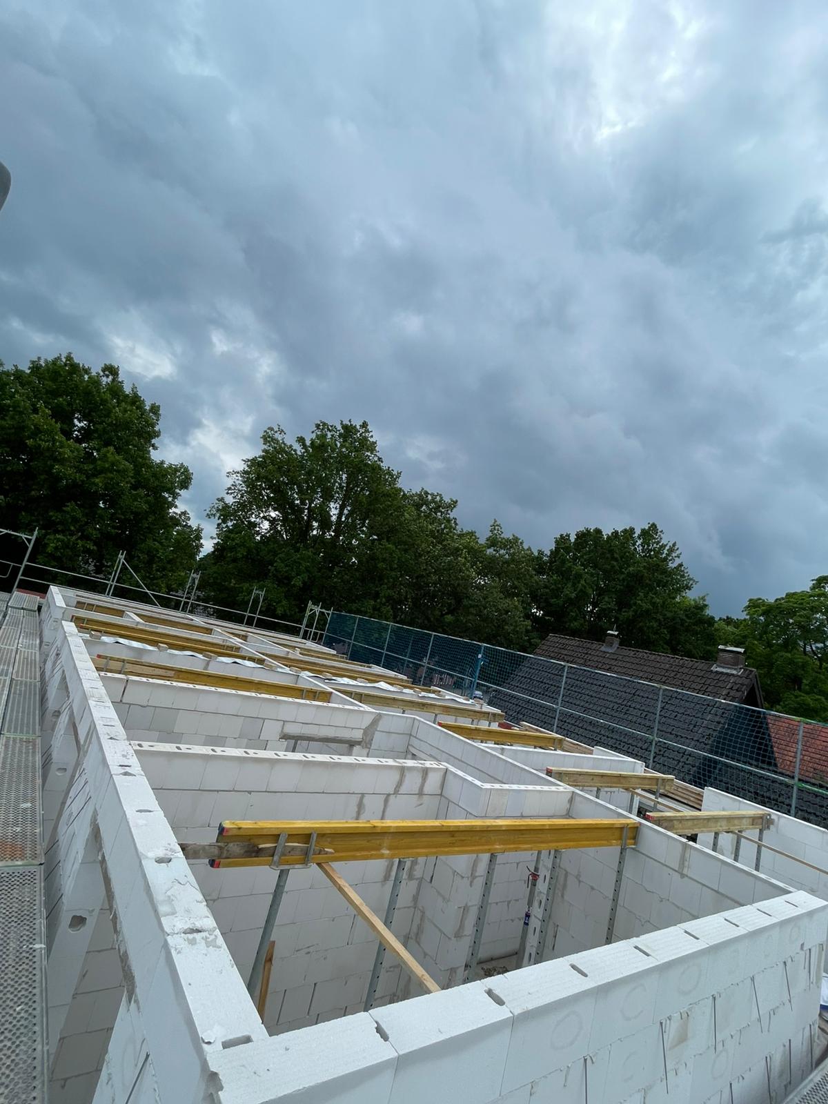 Strukturbau GmbH Oldenburg Teilweise errichtetes Gebäude aus weißen Blöcken mit freiliegenden Holzbalken und Gerüsten, umgeben von Bäumen und Häusern, unter einem bewölkten Himmel.