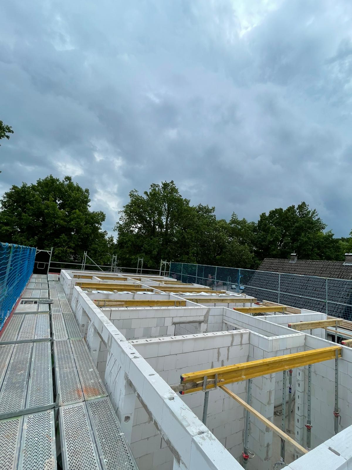Strukturbau GmbH Oldenburg Eine Baustelle mit weißen Betonwänden in Arbeit, gelben Balken, die sich quer über die Baustelle spannen, Metallgerüsten auf der linken Seite und Bäumen und bewölktem Himmel im Hintergrund.