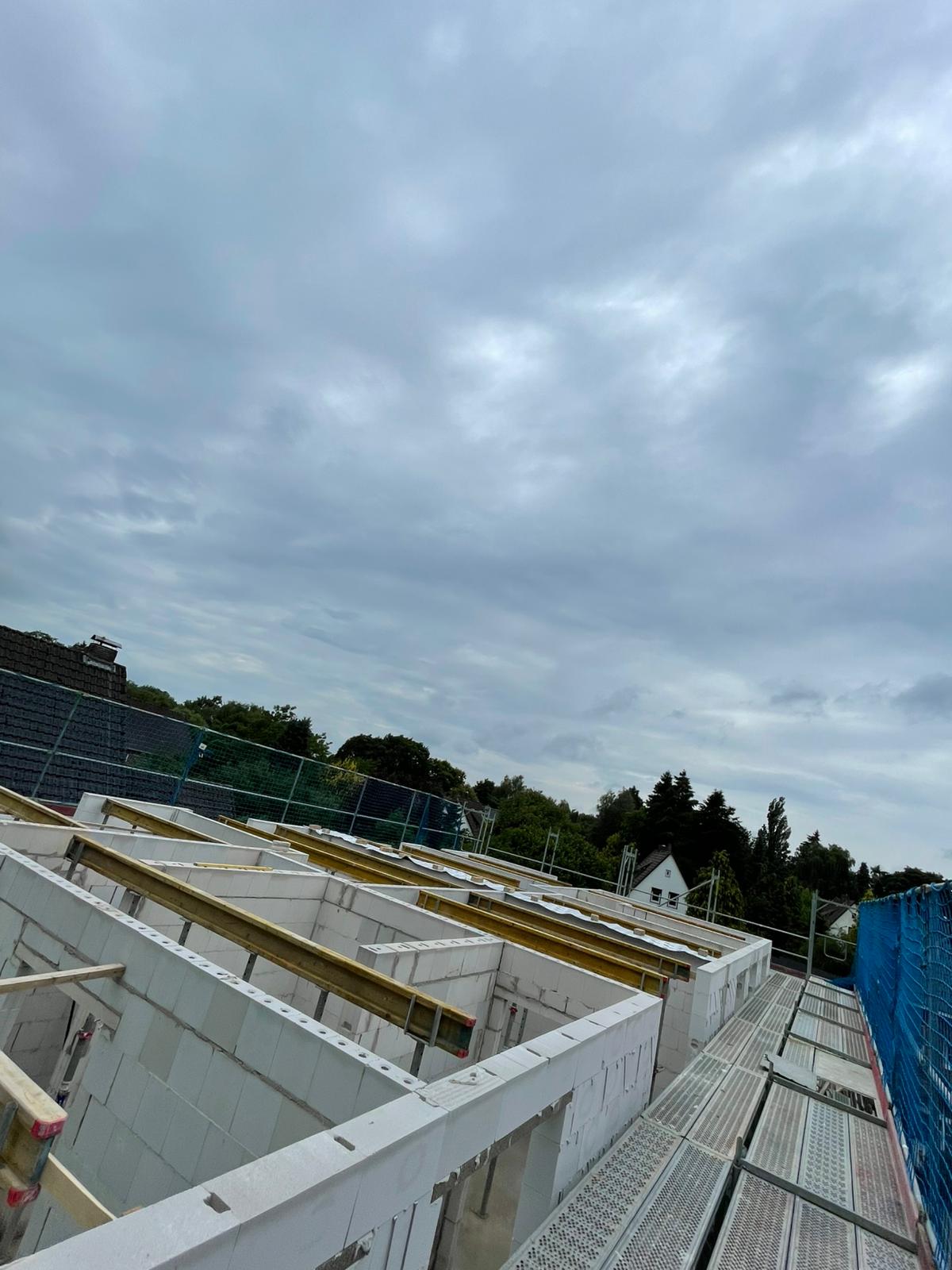 Strukturbau GmbH Oldenburg Eine Baustelle mit teilweise errichteten weißen Betonwänden und freiliegenden Balken, umgeben von blauen Sicherheitsnetzen; im Hintergrund sind Bäume und ein Haus unter einem bewölkten Himmel zu sehen.