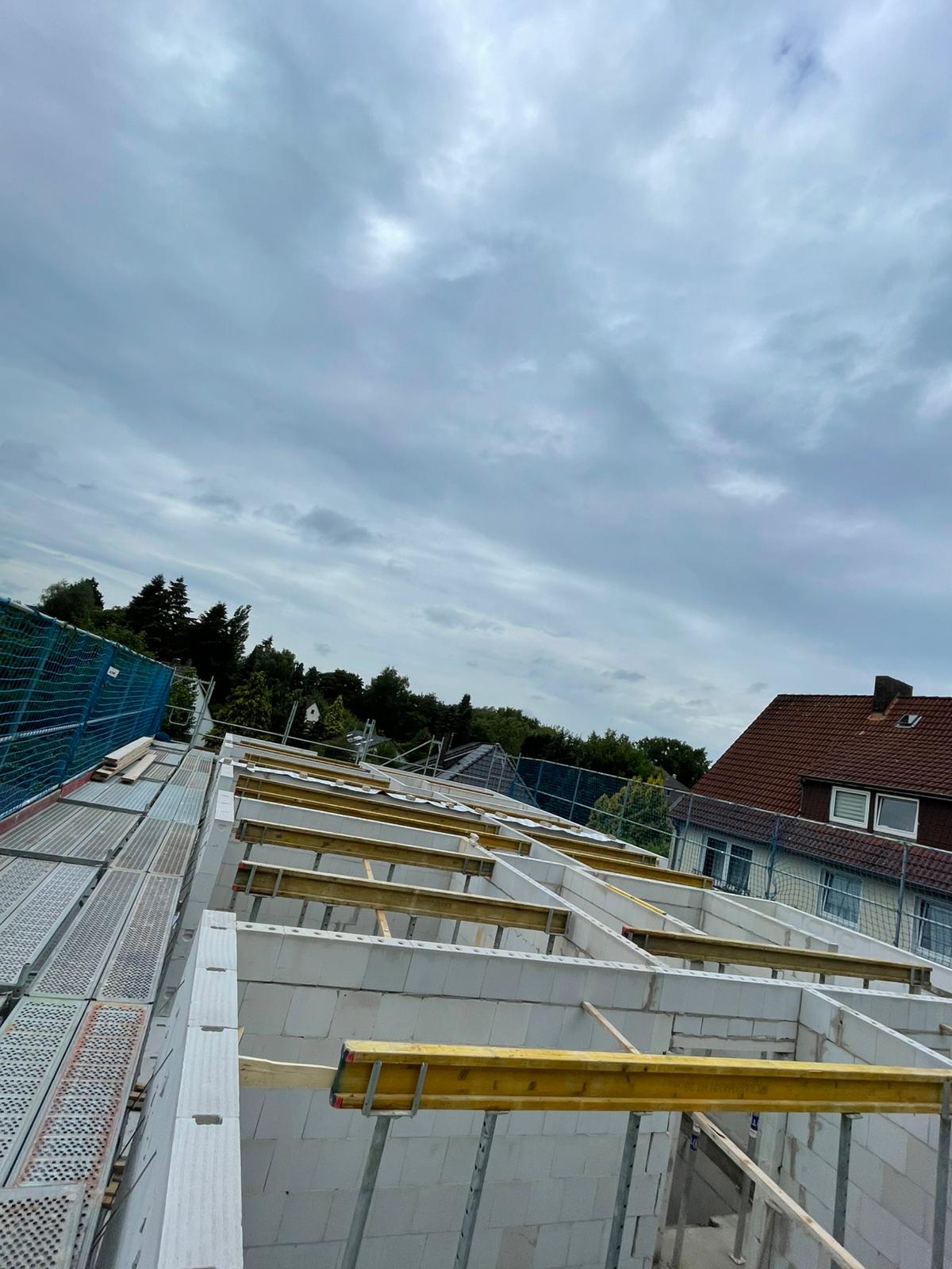 Strukturbau GmbH Oldenburg Eine Baustelle mit teilweise errichteten weißen Ziegelwänden und gelben Holzbalken, umgeben von Gerüsten und Sicherheitsnetzen. Im Hintergrund sind ein Wohnhaus und Bäume unter einem bewölkten Himmel zu sehen.