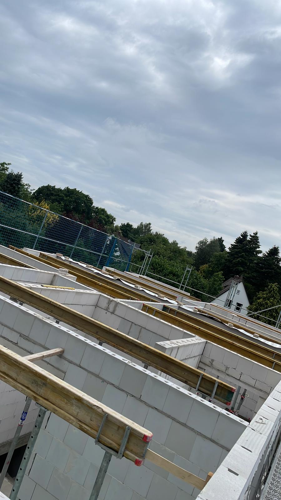Strukturbau GmbH Oldenburg Teilweise errichtetes Gebäude mit weißen Ziegelwänden und Holzbalken, umgeben von Gerüsten und Sicherheitsnetzen. Im Hintergrund sind Bäume und ein Haus unter einem bewölkten Himmel zu sehen.
