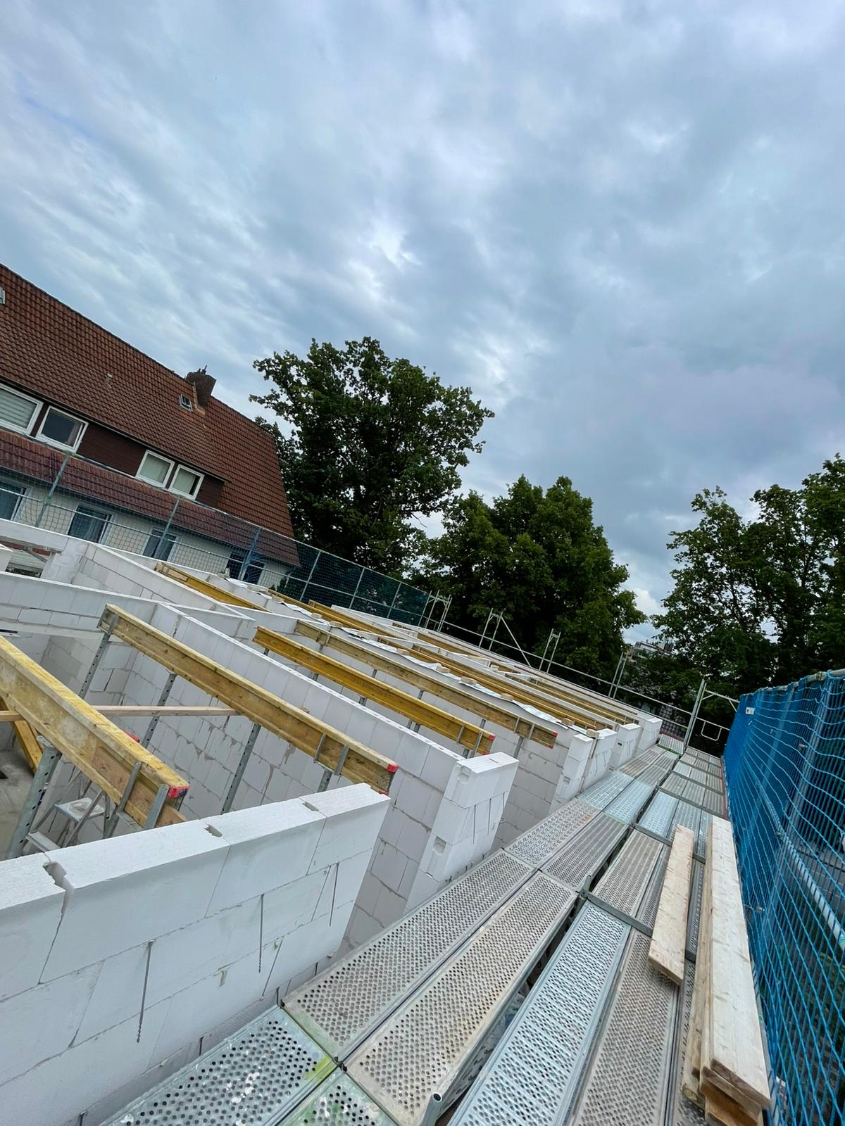 Strukturbau GmbH Oldenburg Eine Baustelle mit teilweise errichteten weißen Ziegelwänden, Holzplanken und Metallgerüsten, neben einem Wohnhaus mit Schrägdach, umgeben von Bäumen unter einem bewölkten Himmel.