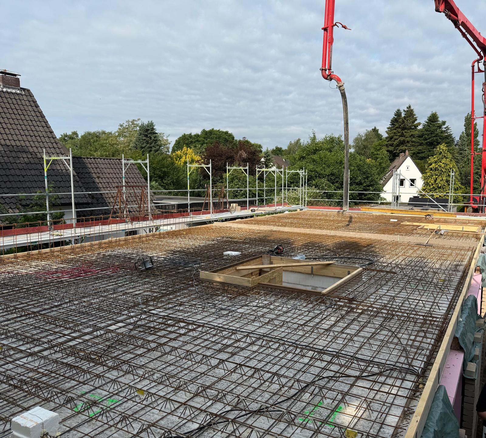 Strukturbau GmbH Oldenburg Baustelle mit einem über ein Betonfundament gelegten Metallgitter, umgeben von einem Baugerüst. Eine Betonpumpe mit einem roten Arm erstreckt sich über die Baustelle, und im Hintergrund sind Bäume und Häuser zu sehen.