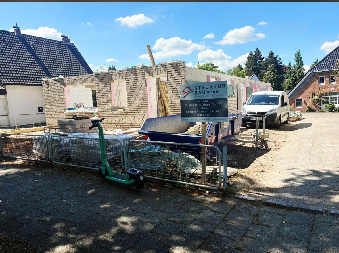 Strukturbau GmbH Oldenburg Ein teilweise errichtetes Backsteingebäude in einer Wohnstraße, mit einem Bauschild, einem weißen Lieferwagen, einem Motorroller und Baumaterialien, die unter einem blauen Himmel mit Wolken verstreut sind.