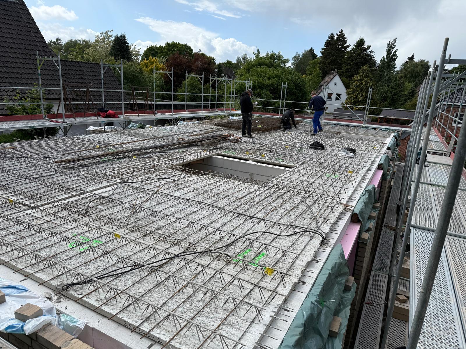 Strukturbau GmbH Oldenburg Bauarbeiter stehen auf einer Baustelle, auf der eine Betonplatte mit Stahlbewehrung verlegt wird, umgeben von Gerüsten, mit Häusern und Bäumen im Hintergrund.