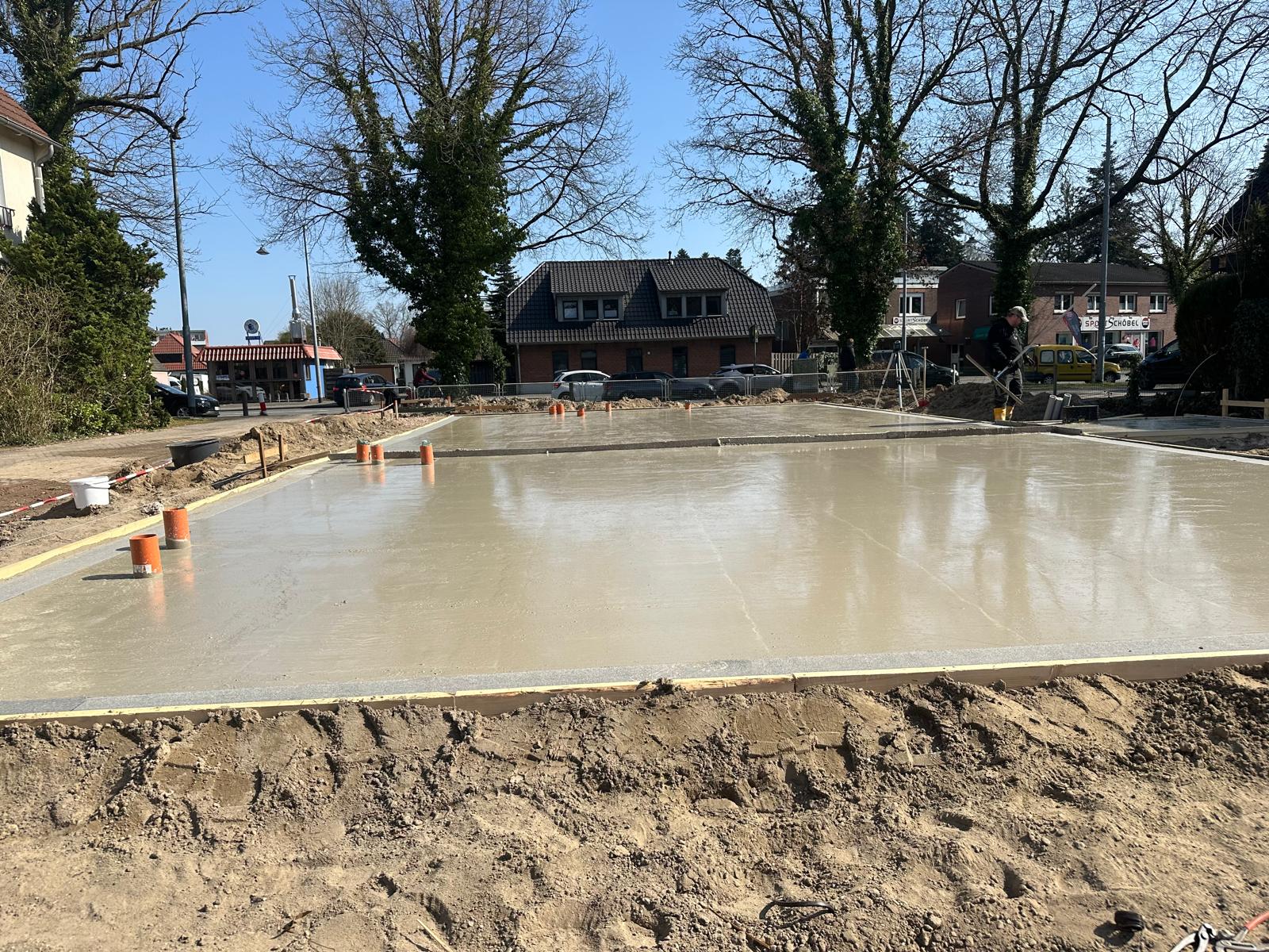 Strukturbau GmbH Oldenburg Eine frisch gegossene Betonfundamentplatte auf einer Baustelle, umgeben von Erde und orangefarbenen Rohren, die aus dem Boden ragen; im Hintergrund sind Wohnhäuser und Bäume unter einem klaren blauen Himmel zu sehen.