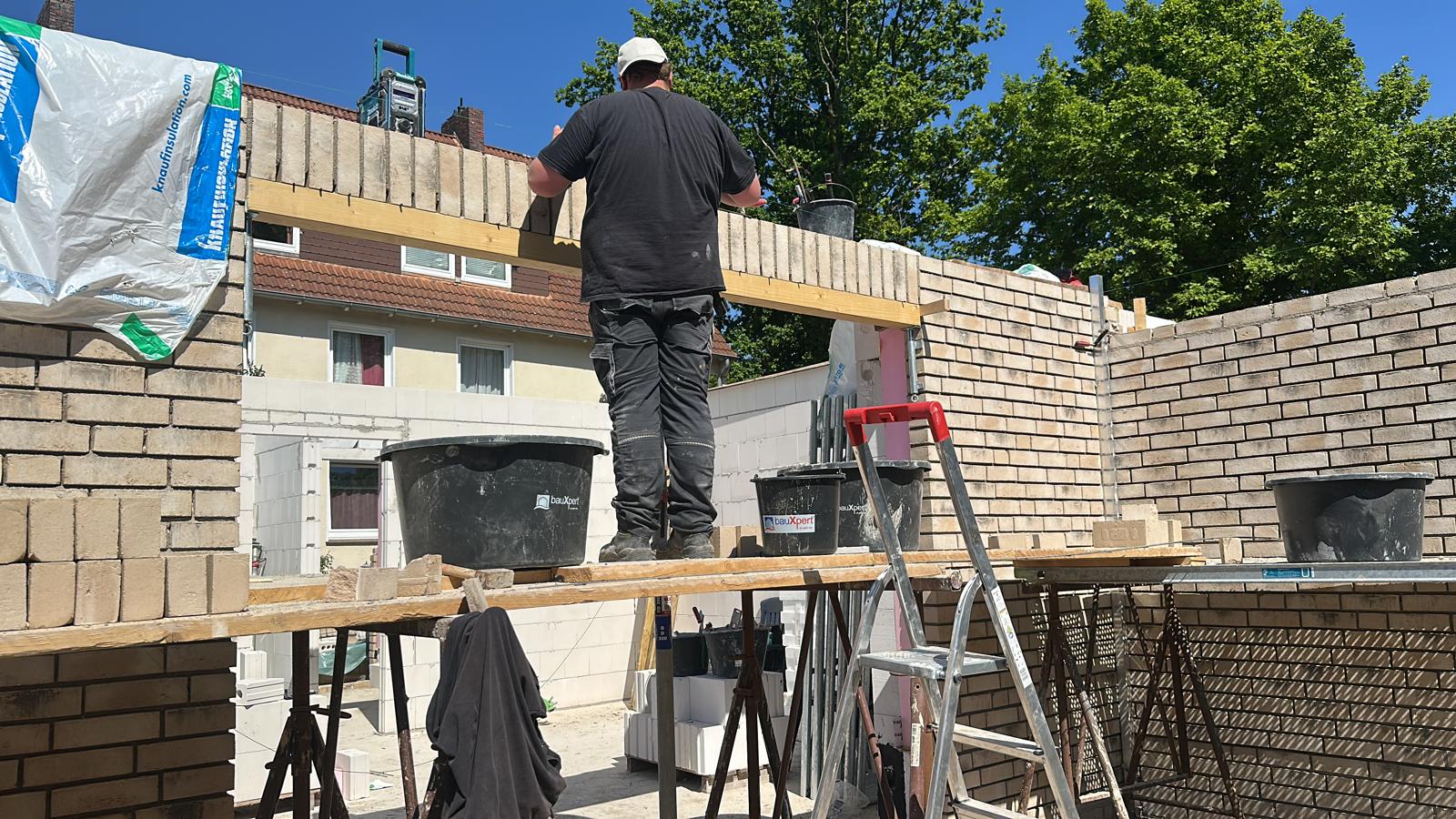 Strukturbau GmbH Oldenburg Ein Bauarbeiter steht auf einem Gerüst und legt unter blauem Himmel Ziegelsteine auf eine teilweise errichtete Hauswand. Eimer und Werkzeuge stehen in der Nähe, und im Hintergrund sind Bäume und Gebäude zu sehen.